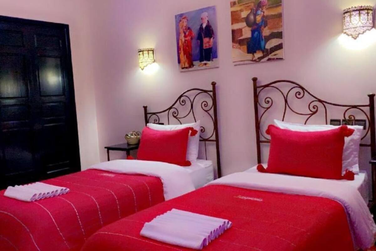 Ein Hotelzimmer mit zwei Betten, beide mit roter Bettwäsche und weißen Kissen. An den Wänden sind Wandleuchten und Kunstwerke über den Betten angebracht.