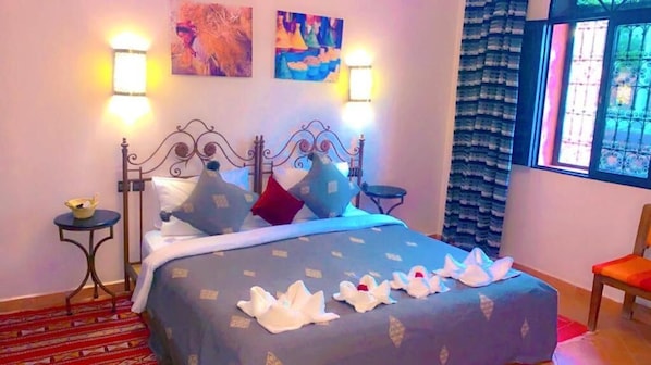 A bed with a blue bedspread, pillows, and decorative swans. There are wall-mounted lights, a window with a view, and a chair.