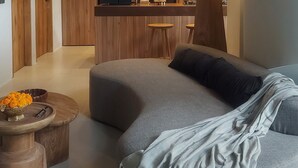 Un salon moderne avec un canapé d’angle gris, une table basse et un coin cuisine doté de meubles en bois et d’un réfrigérateur noir.