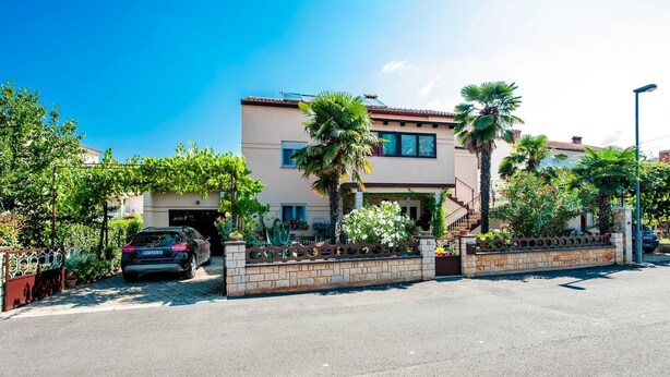 A two-story house with a driveway, a car parked in the driveway, and a well-maintained garden with palm trees and flowering plants. - Apartment in Rovinj near Fantasia Monfiorenzo (Rovinj)