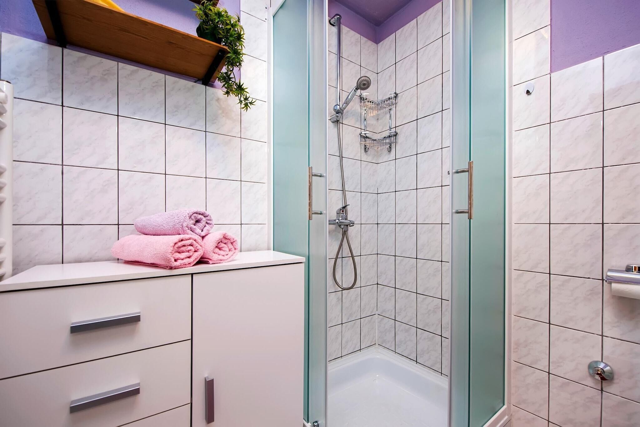 A bathroom with a glass-enclosed shower, white tiled walls, a white sink with a mirror, and a shelf with folded towels.