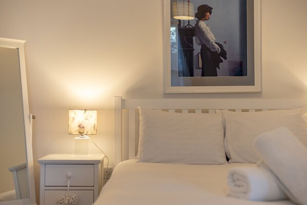 A neatly made bed with white linens and pillows, a bedside table with a lamp, and a framed picture on the wall. - Cosy Garden Cottage | Fleming Place (St Andrews)