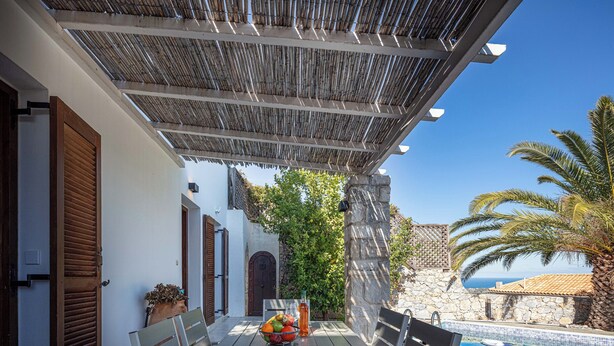 A covered outdoor seating area with a table set for two, a bowl of fruit, and a bottle of water. There is a pool with lounge chairs and a view of the sea. - Villa Delfi /w private pool, at a serene village (Agia Triada)