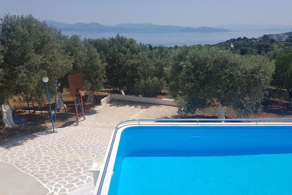 Uma piscina com um caminho de pedra que a leva, rodeada por oliveiras e vista para o mar e montanhas.