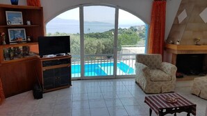 A living room with a TV, a sofa, a small table, and a view of a pool and mountains. - Villa Konaki (Grammatiko)
