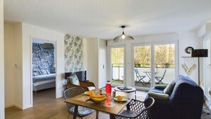 A modern living room with a dining area, a sofa, and a table set with fruits and a red bottle.