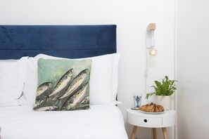 A neatly made bed with a blue headboard, white pillows, and a decorative pillow with fish illustrations. A bedside table with a lamp, a glass of water, a plant, and a croissant is visible. - 3 Pentowan Court -  an apartment that sleeps 2 guests  in 1 bedroom (Carbis Bay)