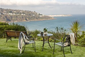 A grassy outdoor seating area with a bench and two chairs, overlooking a coastal landscape with houses and the sea.