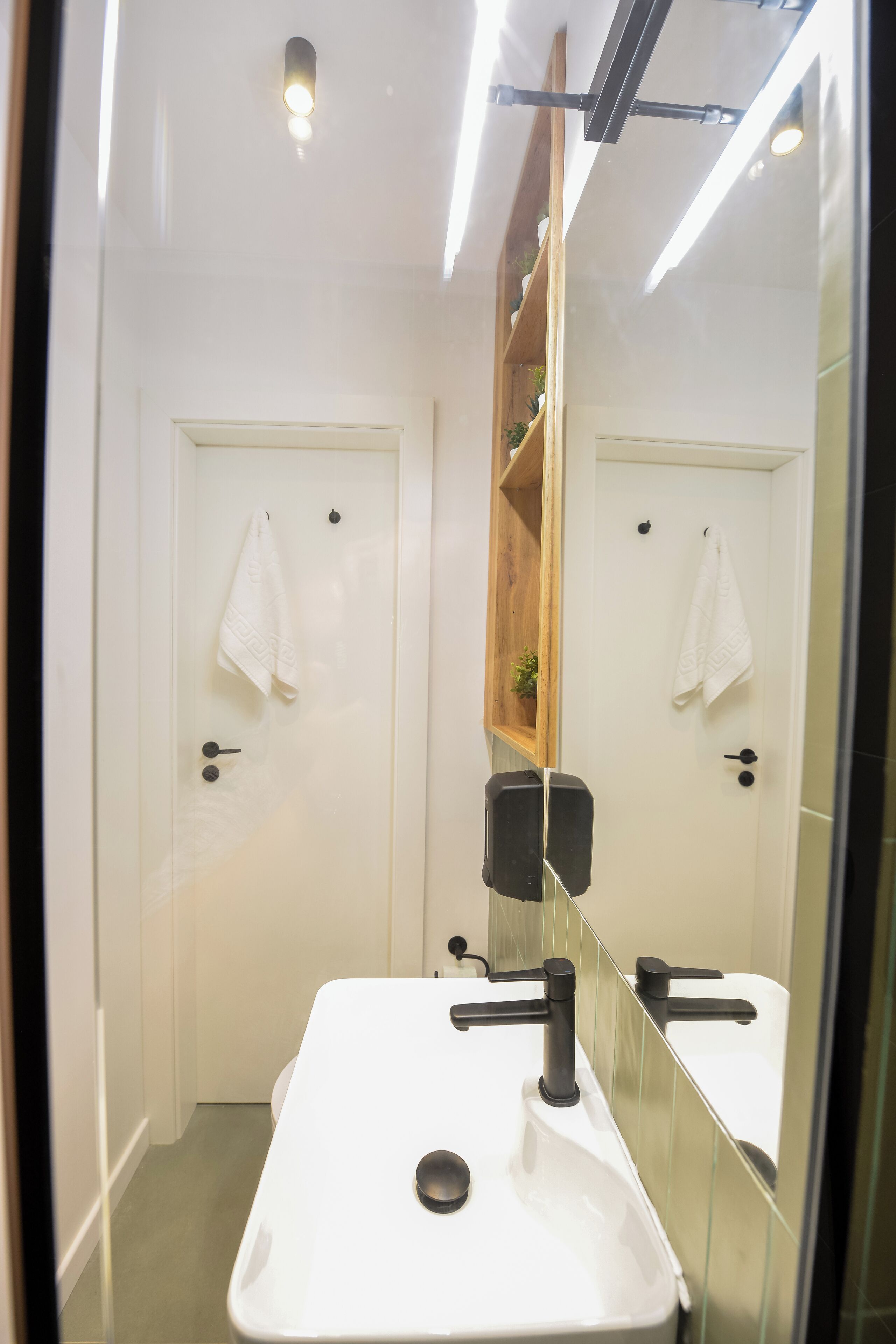Ett modernt badrum med en vit tvättställ, två handdukshängare, en trähylla med en växt och en spegel som reflekterar rummet.