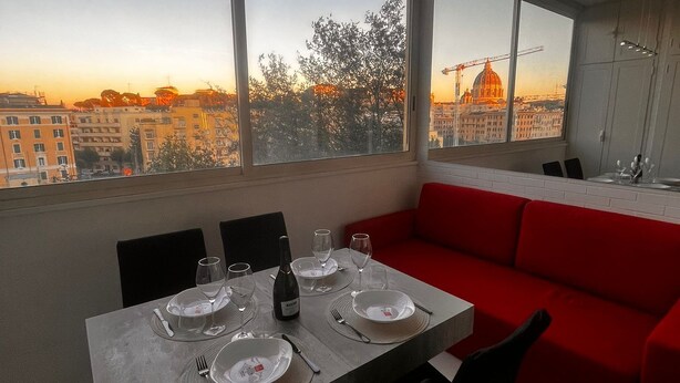 A modern dining area with a table set for four, large windows offering a city view, and a red sofa in the background. - Modern downtown apartment with panoramic view of St. Peter's dome (Roma)