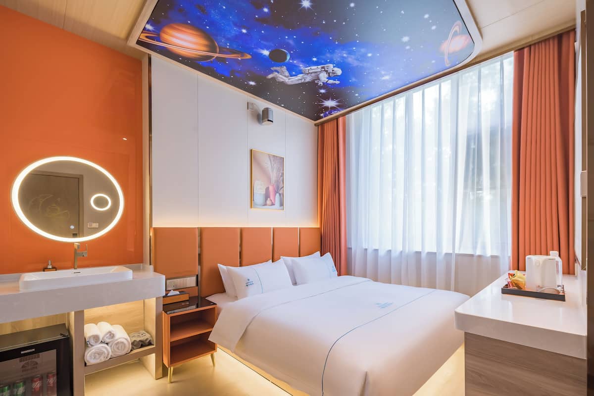 A hotel room with a bed, a bedside table, a mirror, a ceiling mural of space, and a window with curtains.