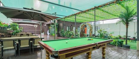 An indoor pool table with a green felt surface, surrounded by wooden furniture and a glass enclosure.