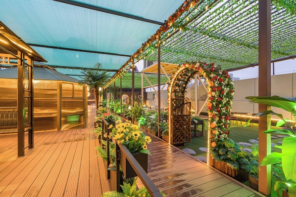 Un espacio al aire libre techado con una terraza de madera, plantas en macetas y un arco floral decorativo.