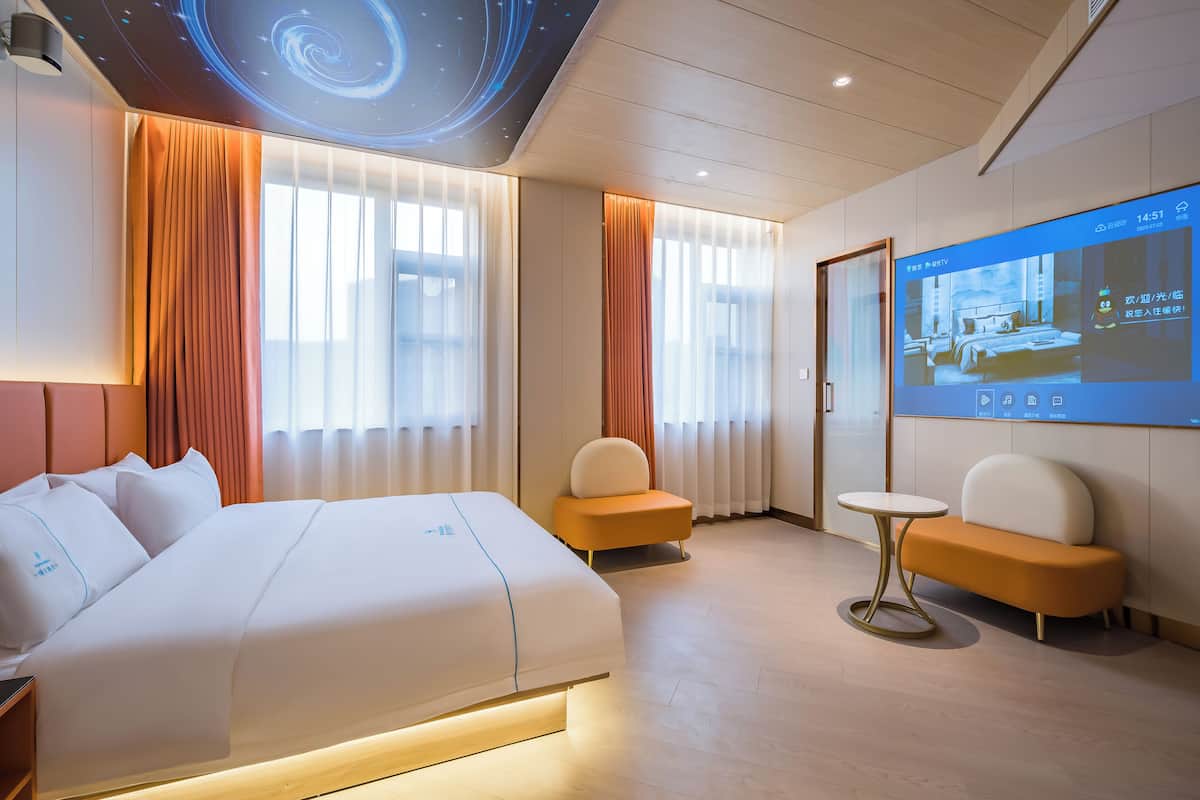 A hotel room with a large bed, two armchairs, a small table, and a wall-mounted TV displaying a room image.
