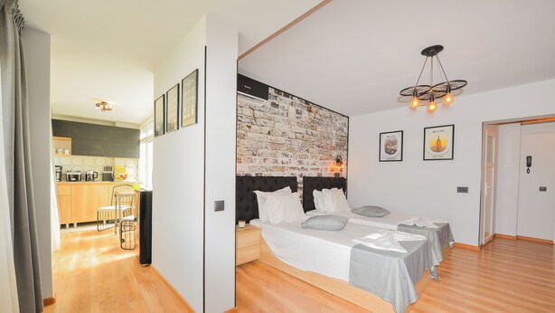 A modern bedroom with a large bed, a small desk, and a kitchen in the background. - One by One - Adam's Studio - Cismigiu Gardens (București)