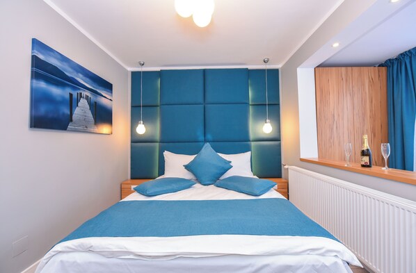 A modern bedroom with a blue headboard, a bed with blue bedding, a nightstand with a bottle and glasses, and a painting on the wall.