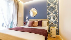 Uma cama bem arrumada com um edredom branco e castanho, um espelho redondo acima e uma mesa de cabeceira com gavetas e uma luminária.