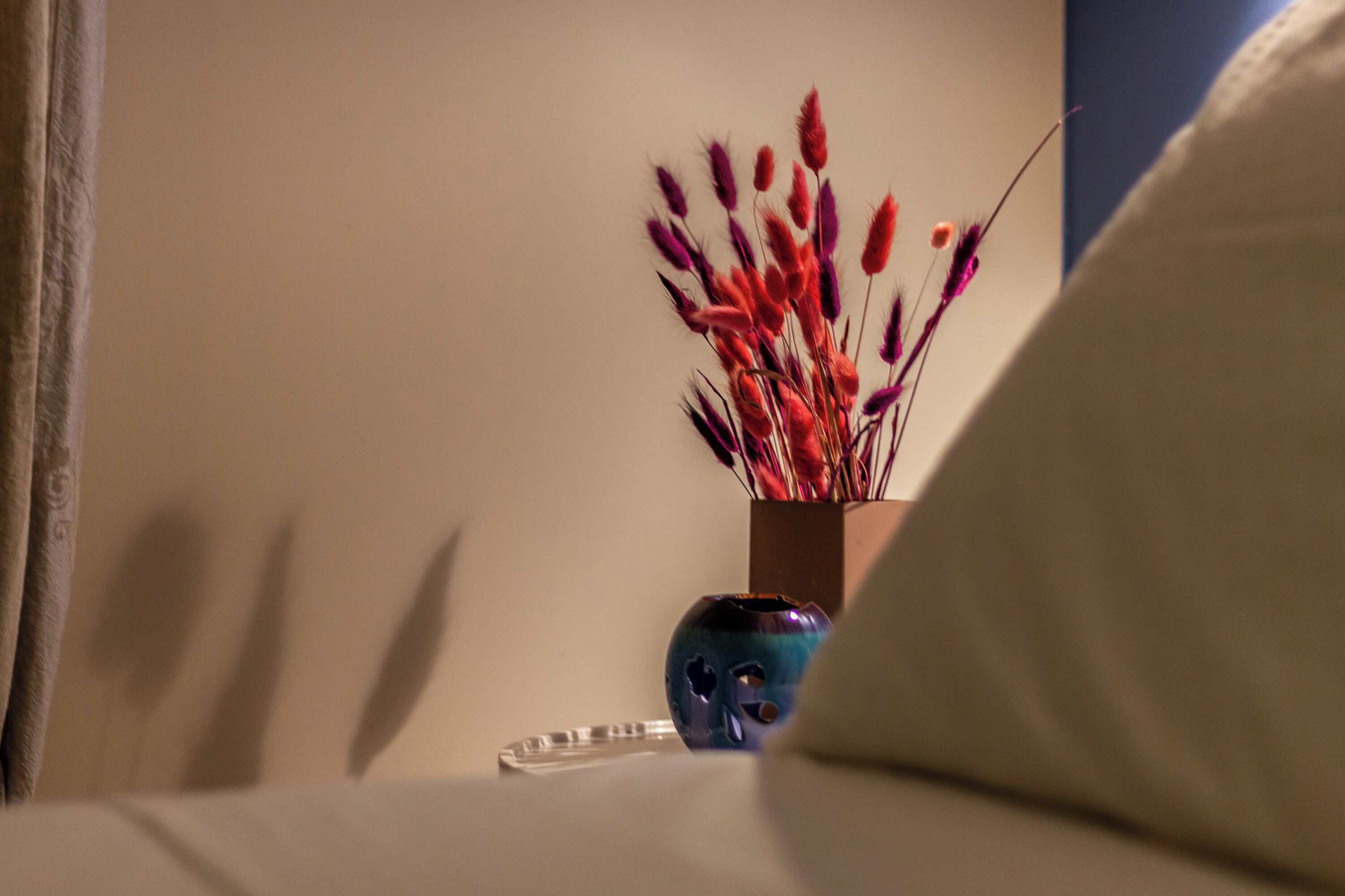Ein Bett mit Kissen, eine Vase mit Trockenblumen und ein kleines blaues Objekt auf dem Nachttisch.