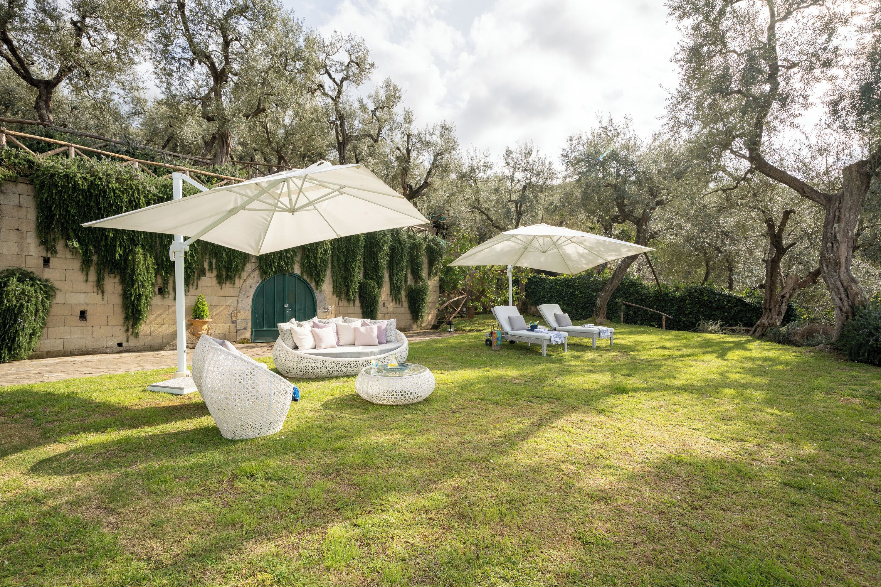 Un jardín con mobiliario exterior blanco, que incluye una zona de estar circular y dos sillones de descanso bajo grandes sombrillas.