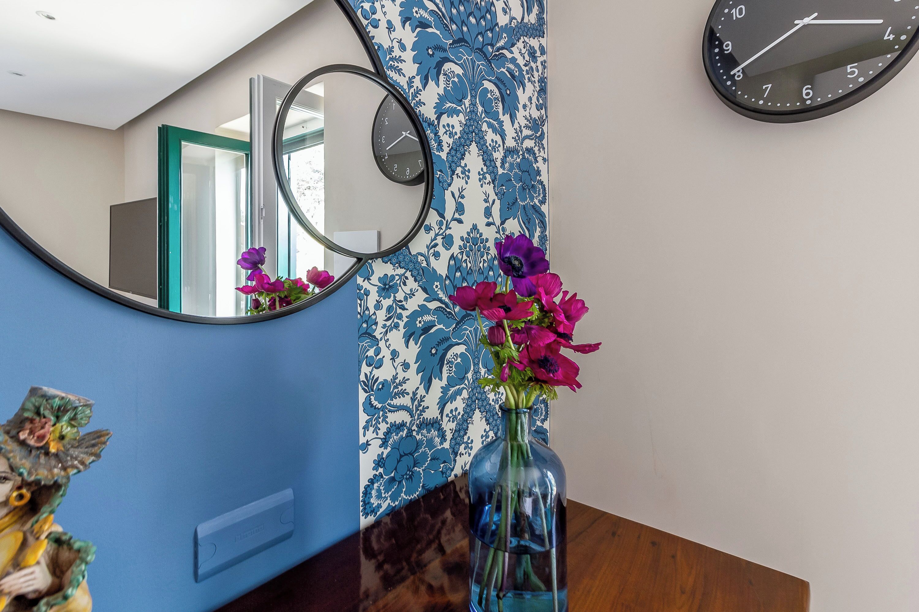Een kamer met blauw-wit behang, een ronde spiegel, een klok en een vaas met bloemen op een houten tafel.
