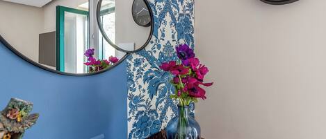 Een kamer met blauw-wit behang, een ronde spiegel, een klok en een vaas met bloemen op een houten tafel.