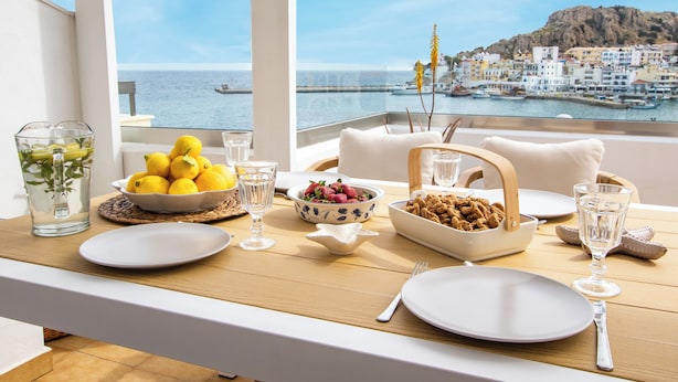 A dining table set with plates, bowls of fruit, and glasses, overlooking a coastal town. - Olia Boutique Apartments (Karpathos)
