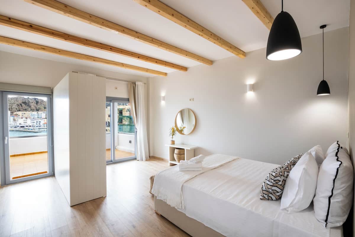 A bedroom with a large bed, wooden beams, a ceiling light, a mirror, and a view of a pool and buildings.