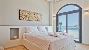 A bedroom with a large bed, a built-in shelf, a decorative wall piece, and a view through a glass door. - Olia Boutique Apartments (Karpathos)