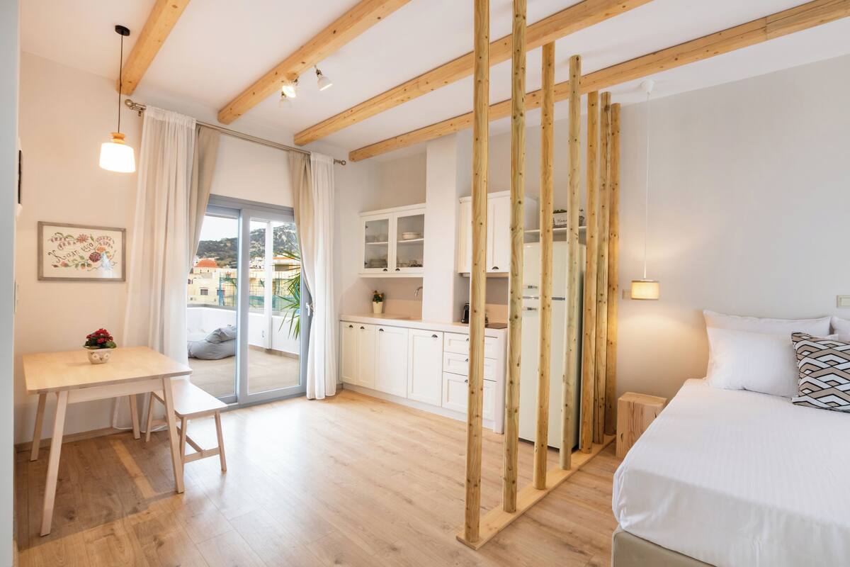 A modern hotel room with a wooden ceiling, a bed with white bedding, a small desk with a chair, a kitchenette, and a sliding glass door leading to a balcony.