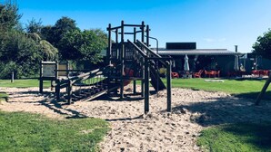 A playground with a climbing structure and a slide, surrounded by sand and grass, with a building and red chairs in the background. - Sunny house close to the dunes and the sea. Families with children and dogs. (Warmenhuizen)