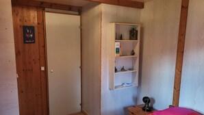 A bedroom with a wooden door, a shelf with books and decorative items, a bedside table with a lamp, and a bed with pink bedding.