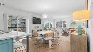 A modern kitchen with a central island, dining area with a round table and chairs, and a living space with a sofa and a TV. - Fun and Sun 2nd Floor Oceanview 3 Bedroom (Pawleys Island)