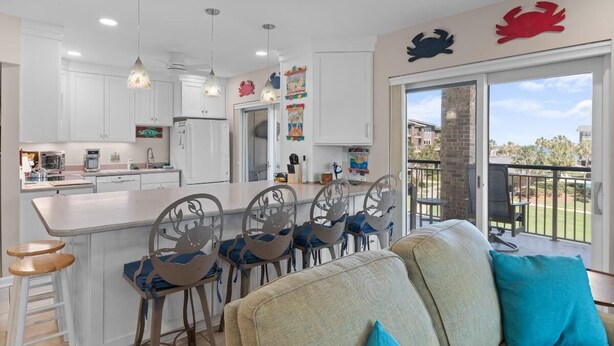 Une cuisine moderne avec des meubles blancs, un îlot central doté de tabourets de bar et un coin repas avec un canapé et des chaises. On y trouve des autocollants décoratifs représentant des crabes sur les murs, et une vue sur l’extérieur grâce à une porte-fenêtre coulissante.