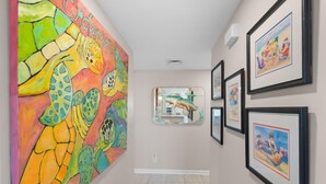 Un couloir orné d’une grande fresque colorée sur le mur de gauche, de tableaux encadrés sur le mur de droite, et d’un miroir qui reflète la pièce.
