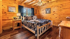 A cozy log cabin bedroom with a wooden bed, a ceiling fan, and a window with curtains. - This cabin is a 3 bedroom(s), 1 bathrooms, located in Sevierville, TN. (Sevierville)