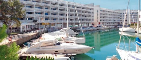 A marina with multiple yachts docked, a white building with balconies, and a clear sky.