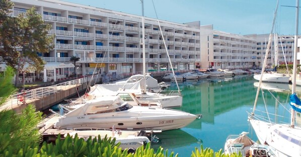 A marina with multiple yachts docked, a white building with balconies, and a clear sky.