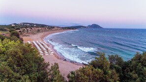 Ein Strand mit Reihen von Sonnenschirmen und Liegestühlen, einer felsigen Küstenlinie und einem entfernten Berg.