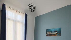 A bedroom with a bed, curtains, a painting, and a hanging light fixture.