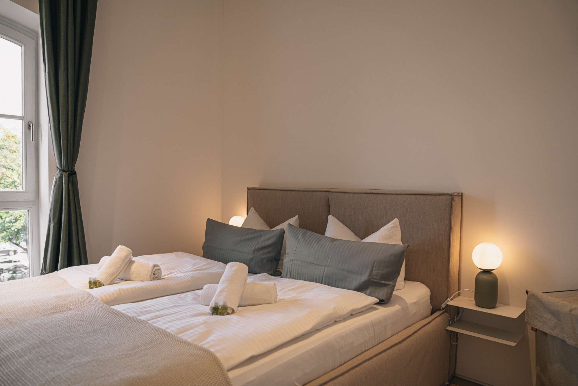 A hotel room with a bed, pillows, a bedside table with a lamp, and a window with curtains.