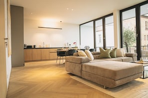 A modern living room with a large sectional sofa, a dining area with a table and chairs, and a kitchen with wooden cabinets.