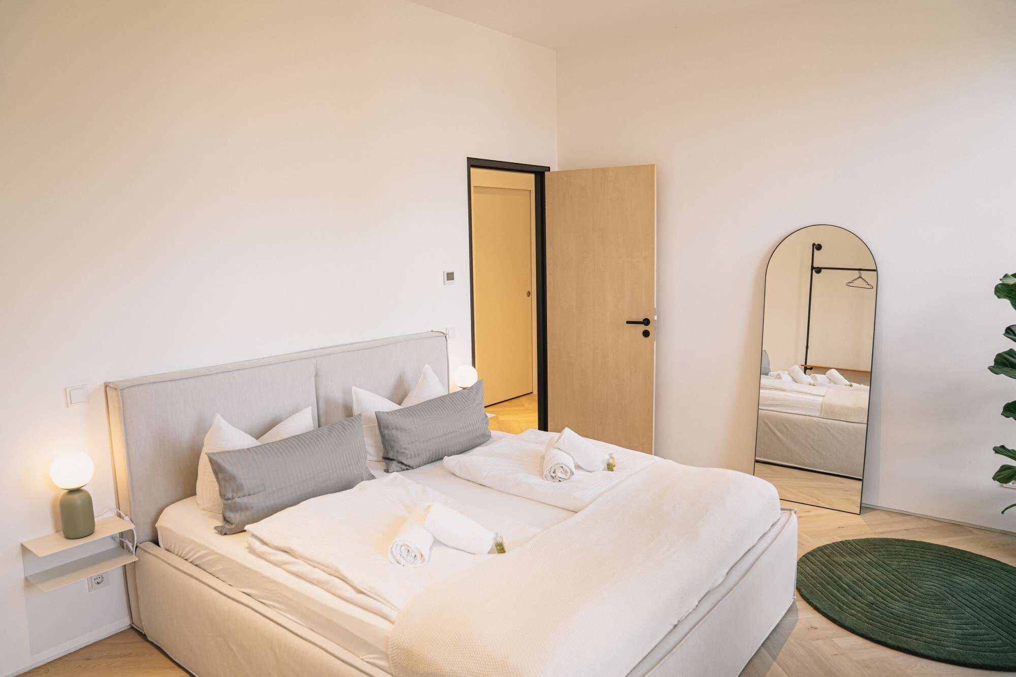 A neatly made bed with pillows and a rolled towel, a bedside lamp, a mirror, and a green rug on a wooden floor.