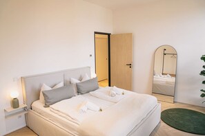 A neatly made bed with pillows and a rolled towel, a bedside lamp, a mirror, and a green rug on a wooden floor.