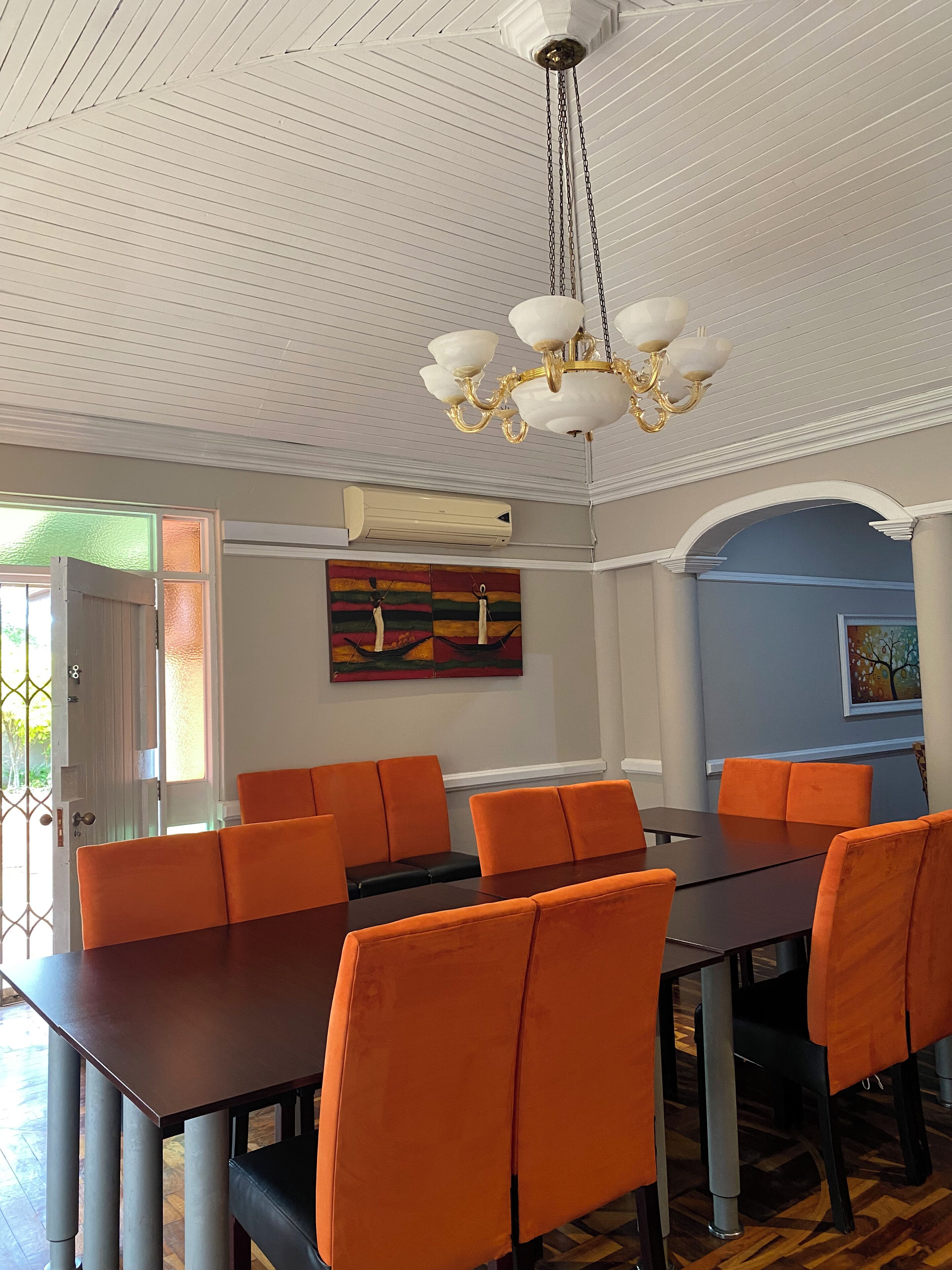 A dining area with a large table and orange chairs, a chandelier, and artwork on the walls.