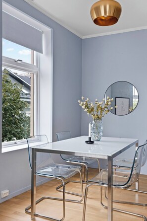 A modern dining area with a clear glass table, metal chairs, a vase with flowers, a round mirror, and a window with a view of greenery.