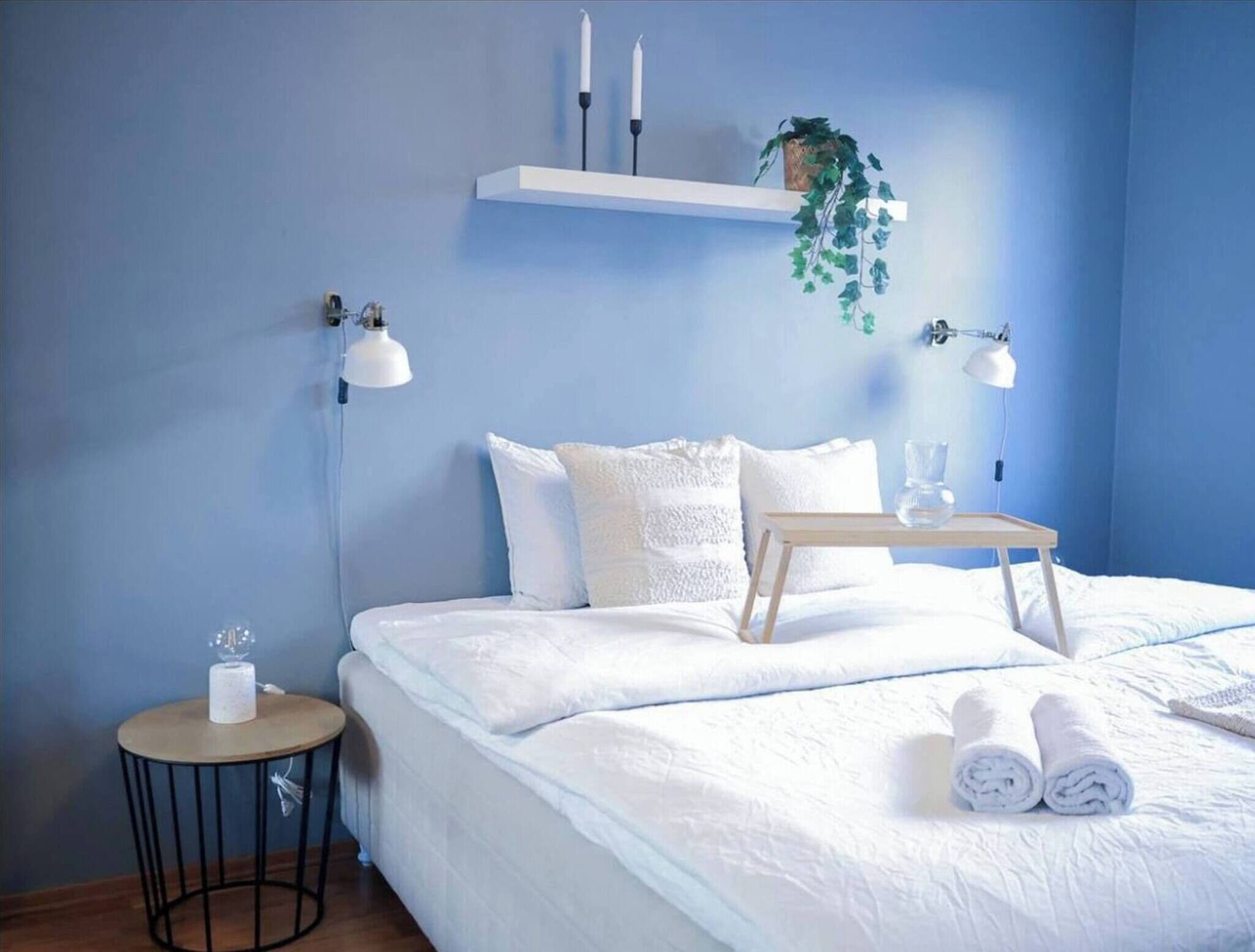 Un dormitorio con una cama, lámparas de noche, una mesita con un vaso y una planta en un estante.