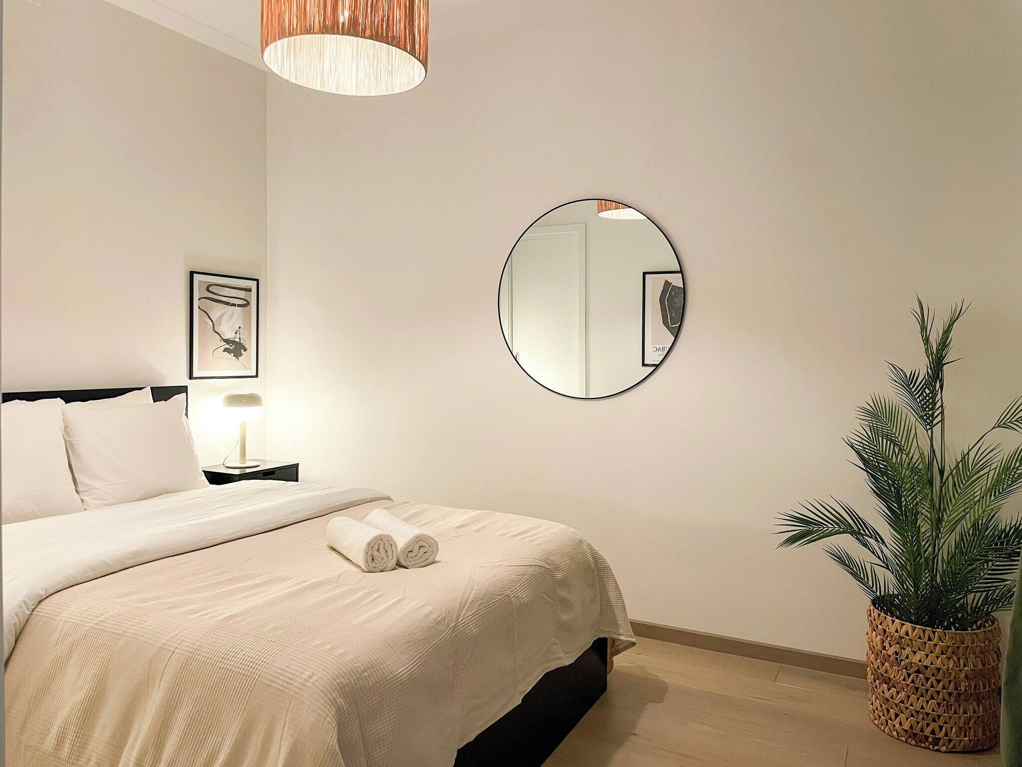 A neatly arranged hotel room with a bed, a round mirror, a bedside lamp, and a potted plant.