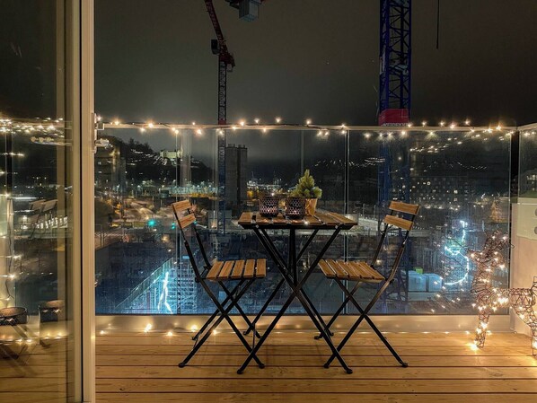 A balcony with a table and chairs, string lights, and a view of a cityscape at night.