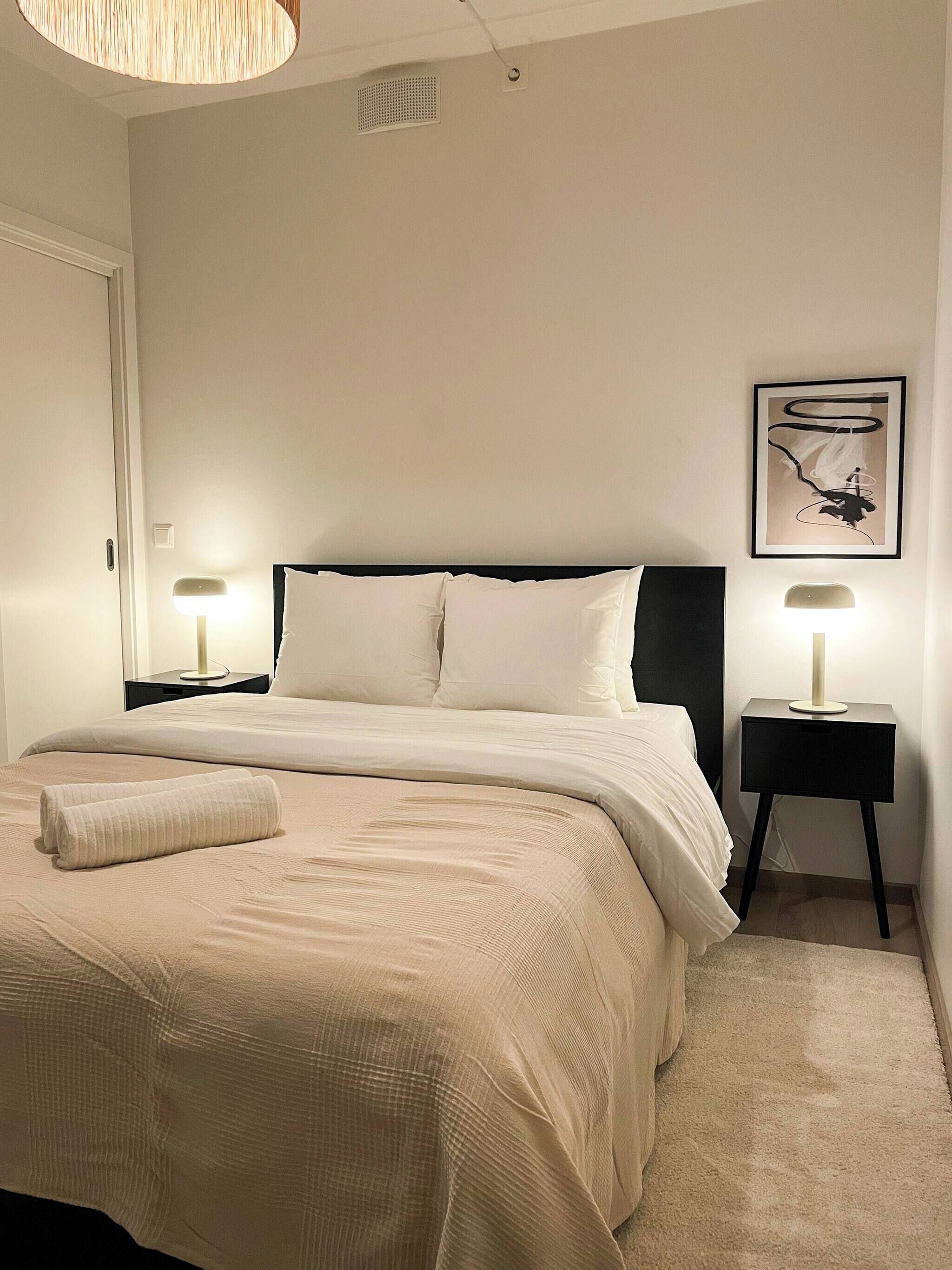 Una cama bien hecha con una toalla enrollada, dos mesitas de noche con lámparas, un cuadro colgado en la pared y una rejilla en el techo.