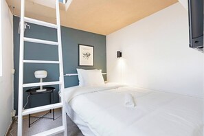 A neatly made bed with white linens, a small bedside table with a lamp, a ladder, and a framed picture on the wall.